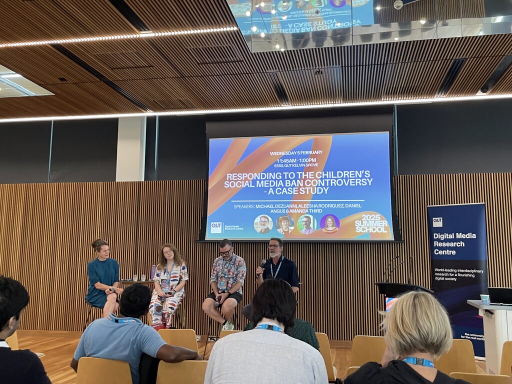 Speakers on a podium in front of a presentation entitled “RESPONDING TO THE CHILDREN'S SOCIAL MEDIA BAN CONTROVERSY - A CASE STUDY”